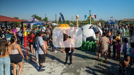 Crowd of families and children enjoying a splash pad with sports-themed water features on a sunny day.