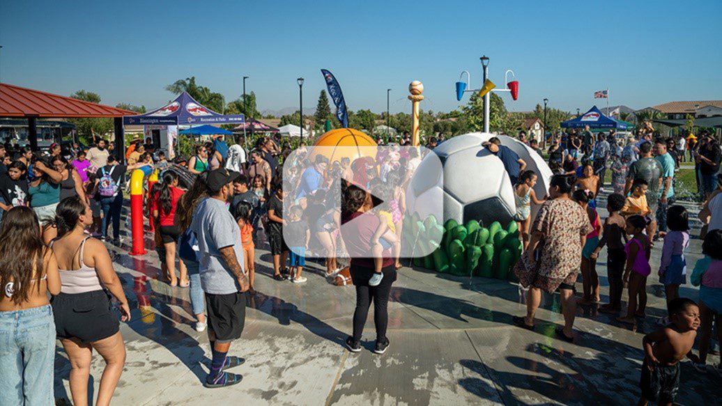 Crowd of families and children enjoying a splash pad with sports-themed water features on a sunny day.