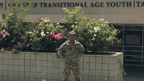 Military man standing in front of bushes and One Stop Transitional Age Youth building.