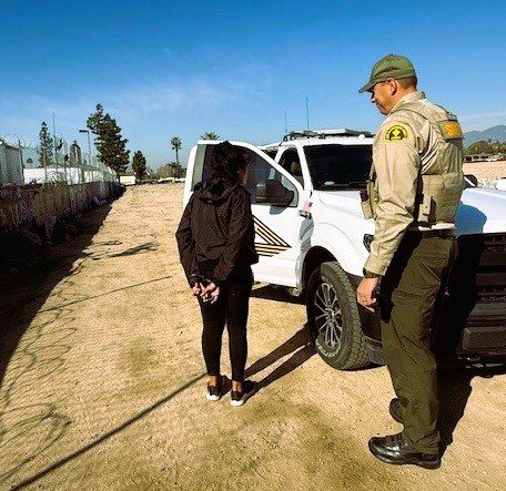 A person is standing with hands behind their back next to a law enforcement officer in uniform. They are beside a white police vehicle parked on a dirt road.