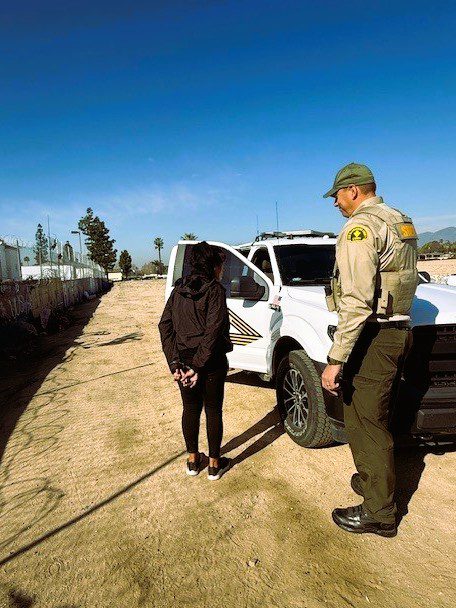 A person is standing with hands behind their back next to a law enforcement officer in uniform. They are beside a white police vehicle parked on a dirt road.