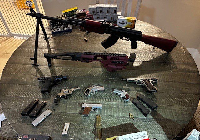 A round wooden table displays various firearms, including rifles, pistols and revolvers, alongside ammunition boxes.