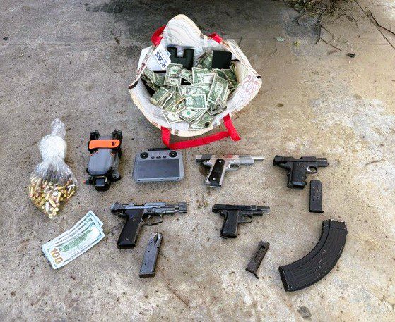 A pile of cash in a bag with binoculars, a clear bag with bullets, a drone and drone controller, and three disassembled guns with three one-hundred-dollar bills on a concrete floor.