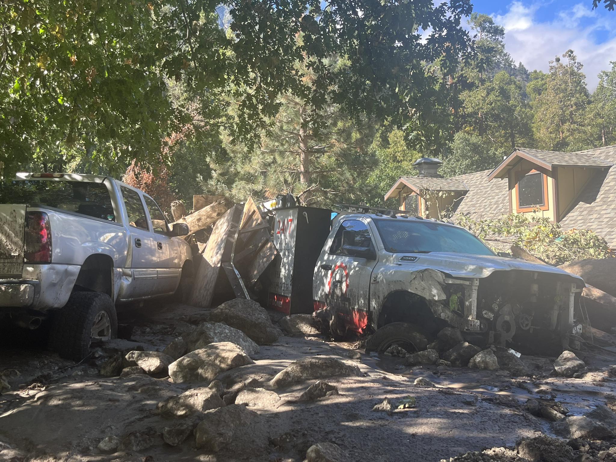 Two damaged pickup trucks sit amid thick mud, fallen branches, and debris in front of a house after a mud and debris flow in a wooded area.