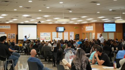 A large group of people sitting in a meeting room listening to a speaker presenting at the front near a projector screen.