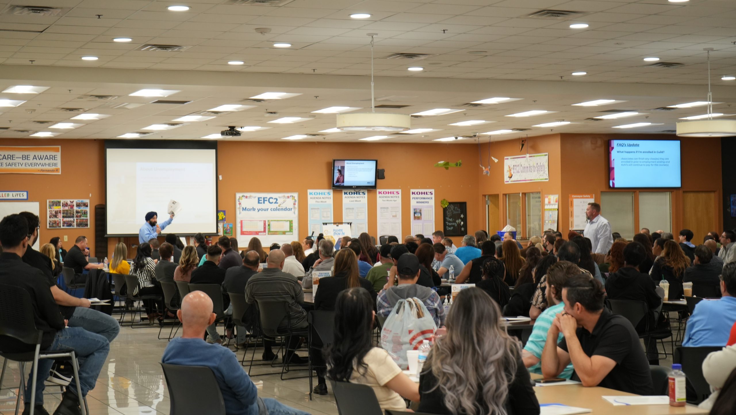 A large group of people sitting in a meeting room listening to a speaker presenting at the front near a projector screen.