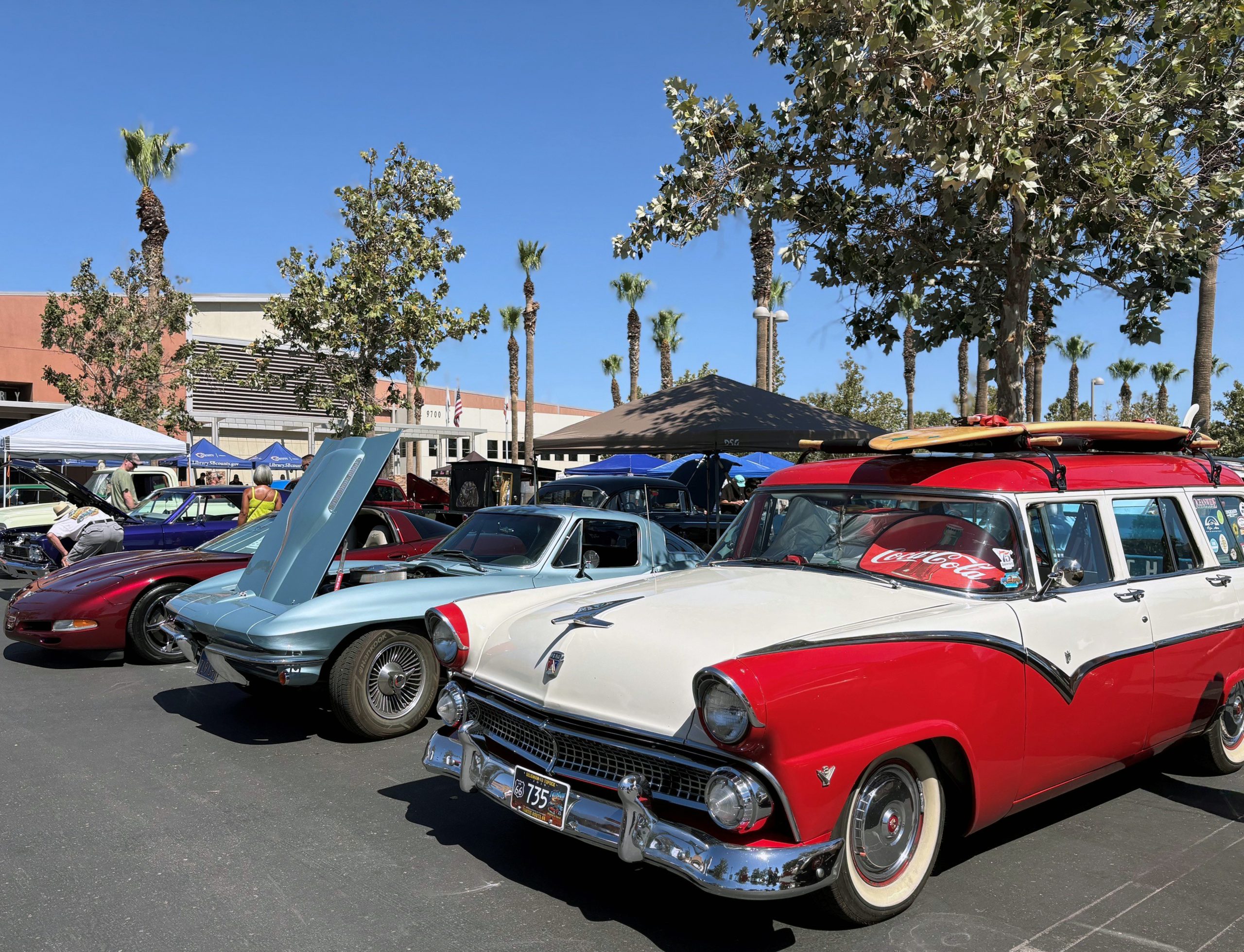 Sunny outdoor classic car show featuring a red-and-white vintage station wagon, polished cars, palm trees, and a lively crowd.
